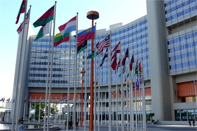[ai] A variety of national flags displayed on poles outside a modern building with large glass windows. The building's design features multiple stories and an orange accent.